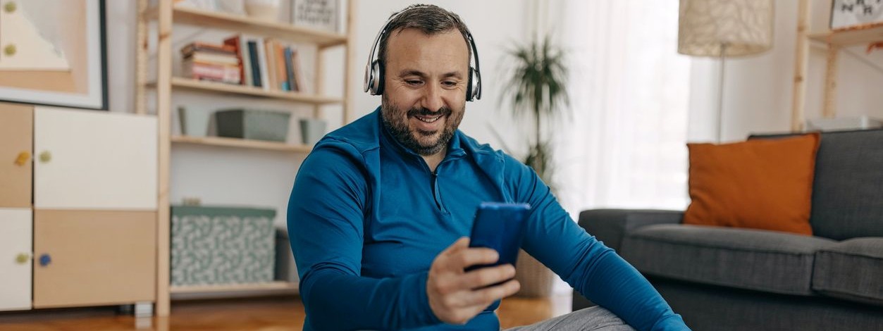 Kräftiger Mann sitzt auf einer Yogamatte und schaut auf sein Handy. Er hat Kopfhörer auf.